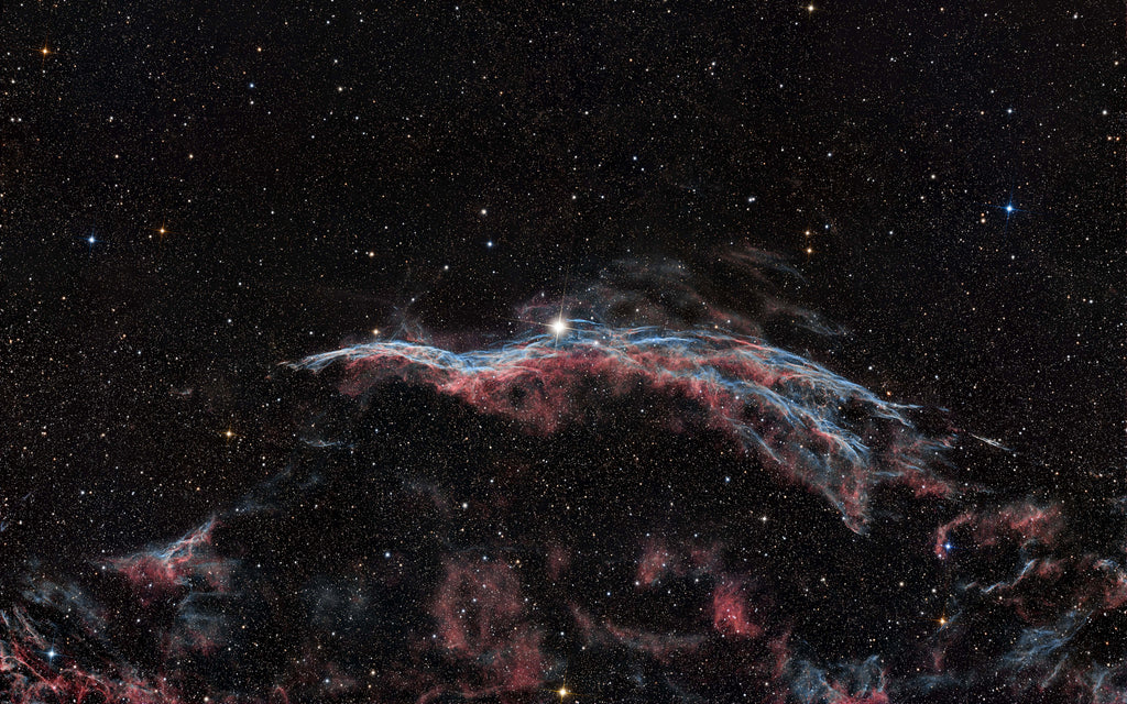 A star-filled deep space scene with a glowing nebula in red, pink, and blue tones against a black background.
