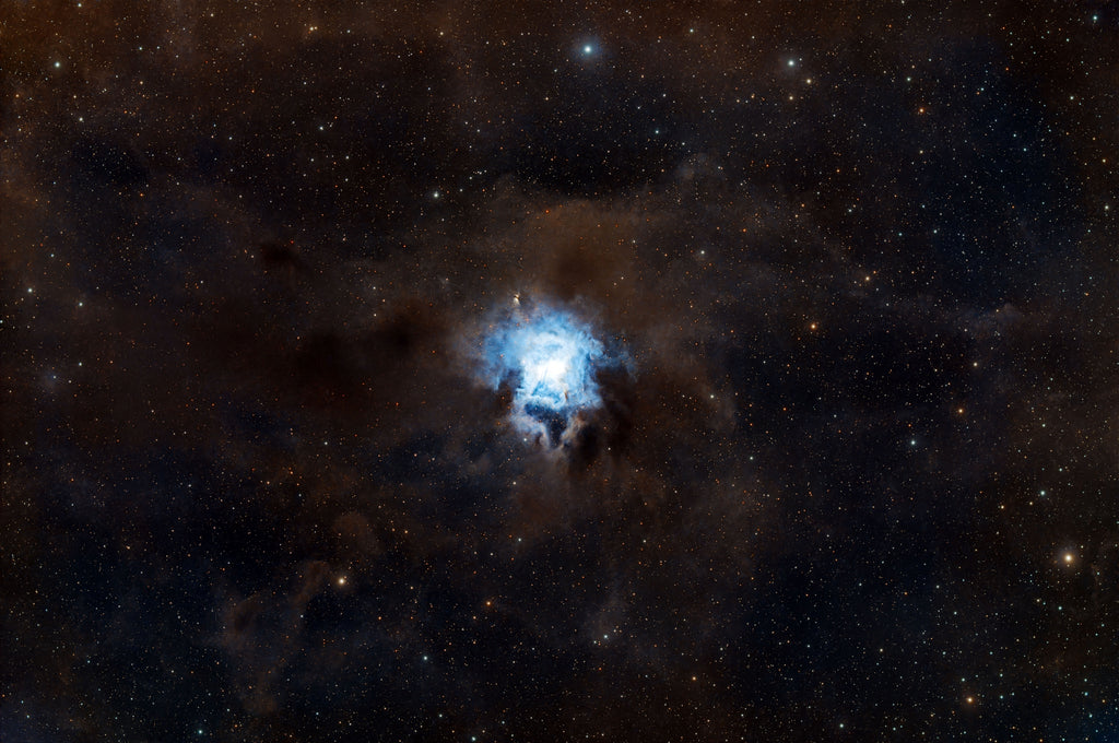 A deep space starfield photograph featuring a bright blue nebula or stellar glow in the center, surrounded by dark brown and black cosmic clouds and numerous small stars.